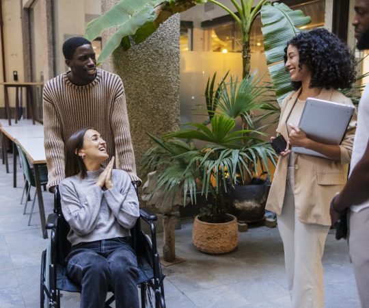 In a vibrant coworking environment adorned with lush plants, a diverse team of professionals engages in discussion, showcasing inclusion and collaboration with a team member using a wheelchair
