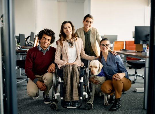 Portrait of happy colleagues with assistance Labrador Retriever dog in the office looking at camera. One of them is in a wheelchair.