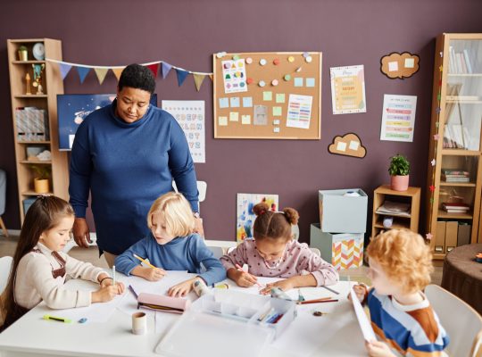 Mature African American nursery teacher standing by table where group of intercultural little learners drawing with crayons at lesson