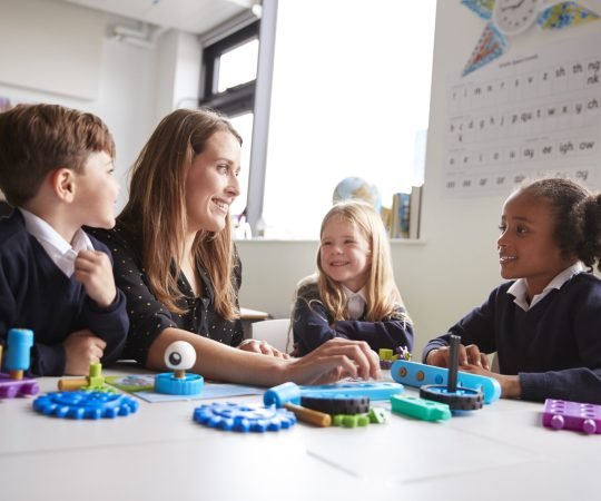 Female teacher and three primary school kids sitting at a table in a classroom working with educational construction toys, close up, low angle